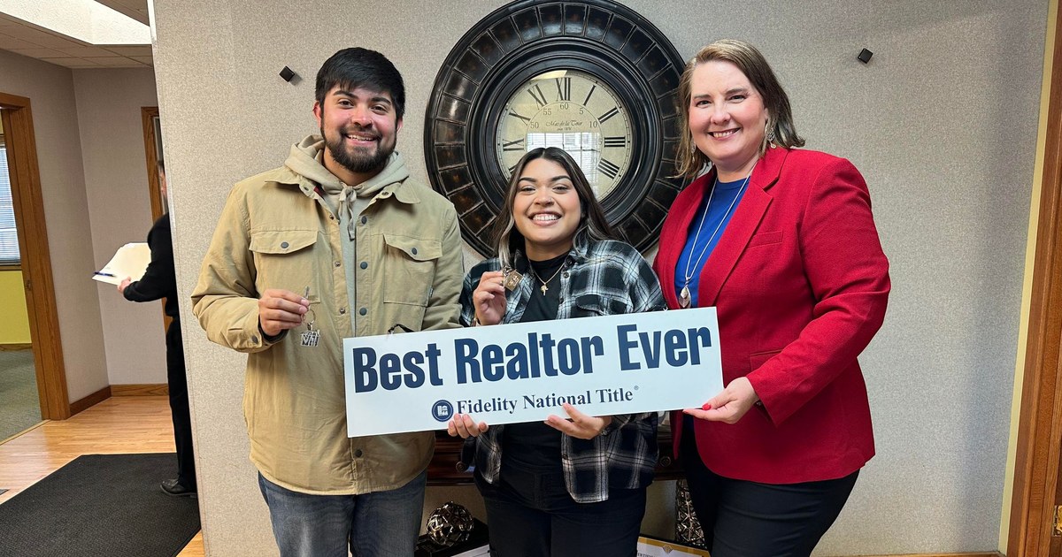 Two new homeowners hold up their house keys beside Lisa Collio, who wears a red blazer, at a title office.