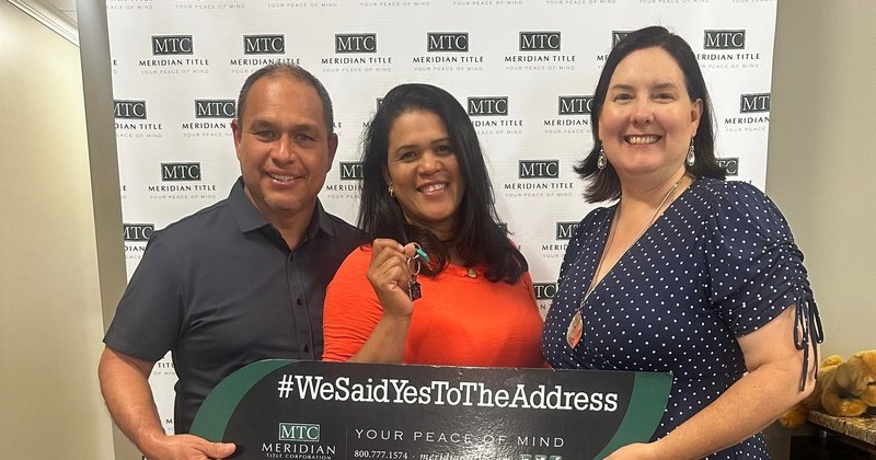 Lisa Collio stands beside a smiling couple at a title office. The woman holds up a house key and the couple holds a long title-company sign.