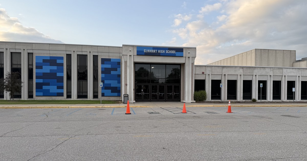 A long single-storey school building with a covered main entrance and traffic cones in the empty lot in front of it, seen under a partly clouded sky.