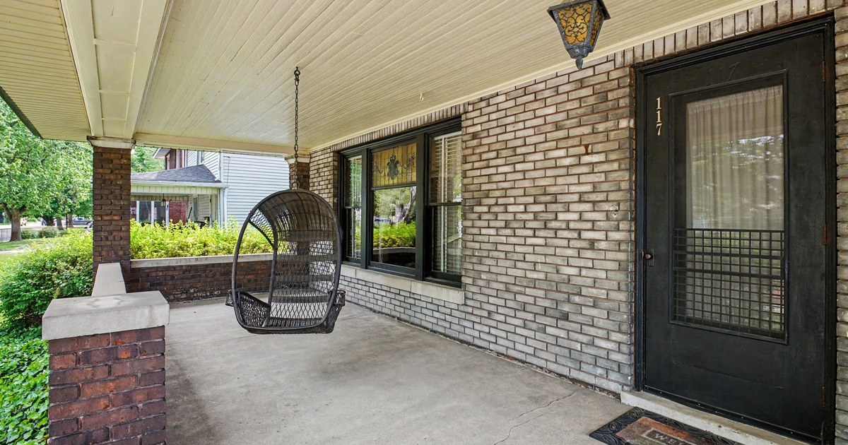 A covered brick front porch with a hanging egg chair beside a dark front door.