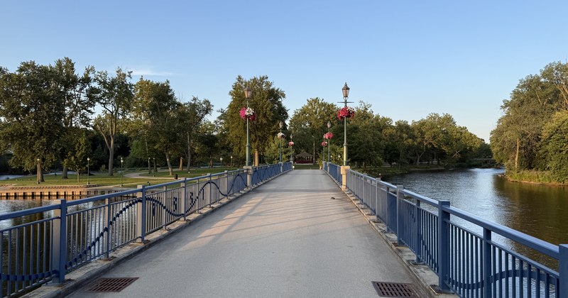 Un puente azul de acero sobre un río que lleva a un parque, con faroles y canastas colgantes de flores a lo largo del barandal.