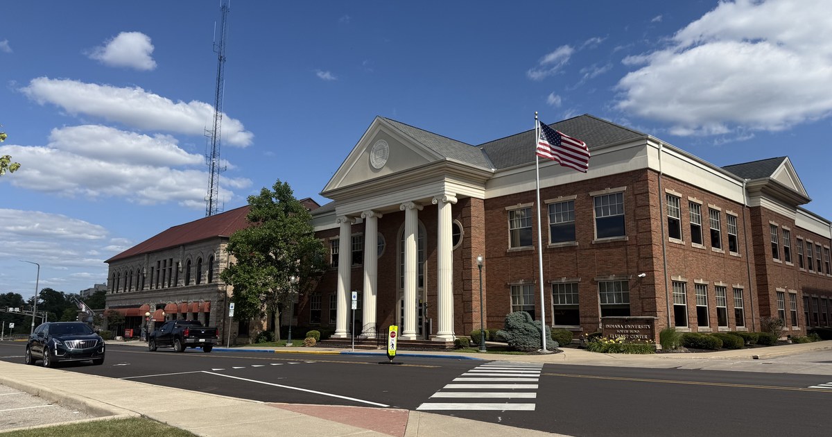 A two-storey brick building with a white columned entrance and a United States flag on a pole, seen from a street corner.