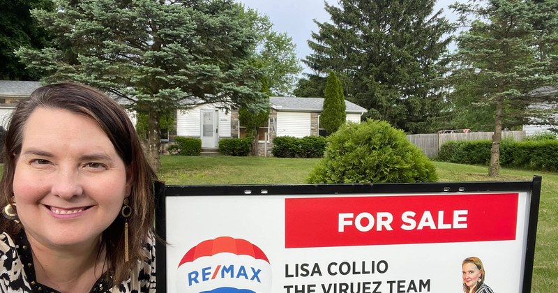 Lisa Collio standing beside a RE/MAX for-sale sign outside a home in summer.