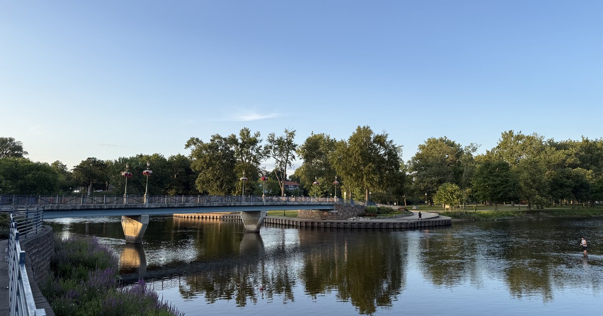 A river passing a low bridge and a wooded park bank in evening light.
