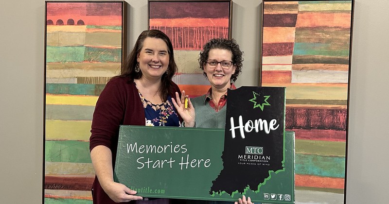 Lisa Collio and a woman stand side by side at a title-office table, holding an Indiana-shaped home sign and a house key.