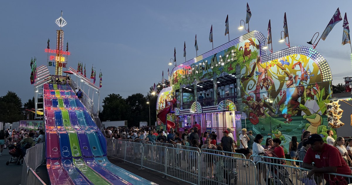 A fairground midway at dusk, with a brightly lit multi-lane slide on the left and a painted funhouse ride under strings of lights on the right.