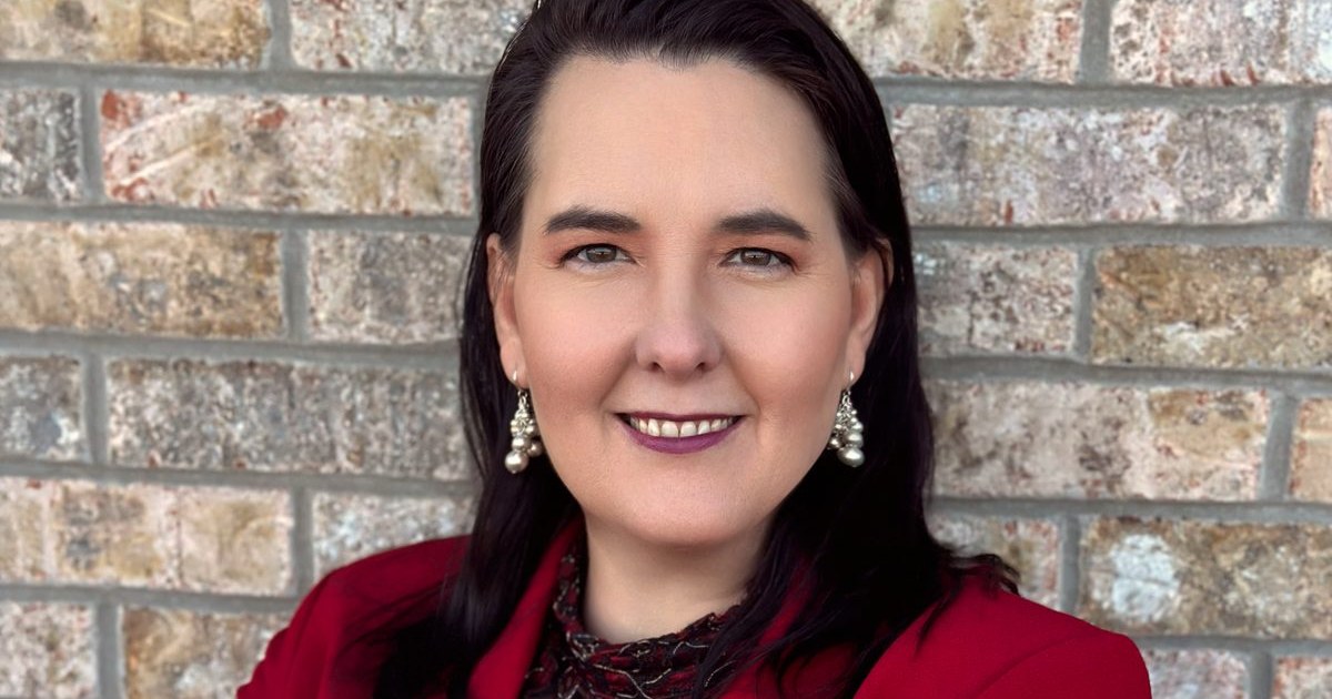 Lisa Collio in a red blazer standing by a brick wall.