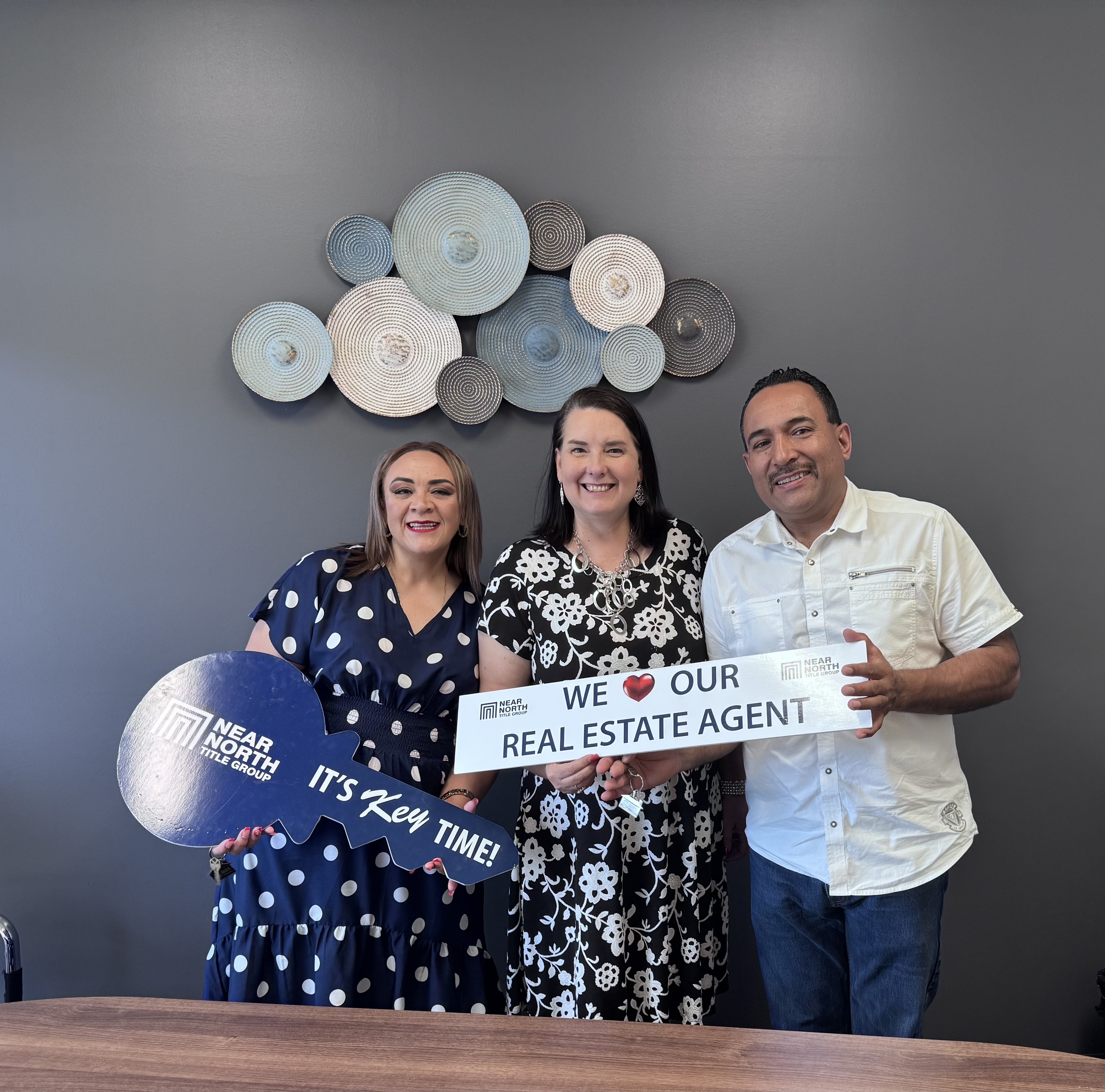Lisa Collio y un cliente de pie en interiores, sosteniendo un letrero rojo en forma de casa que dice "Sold".