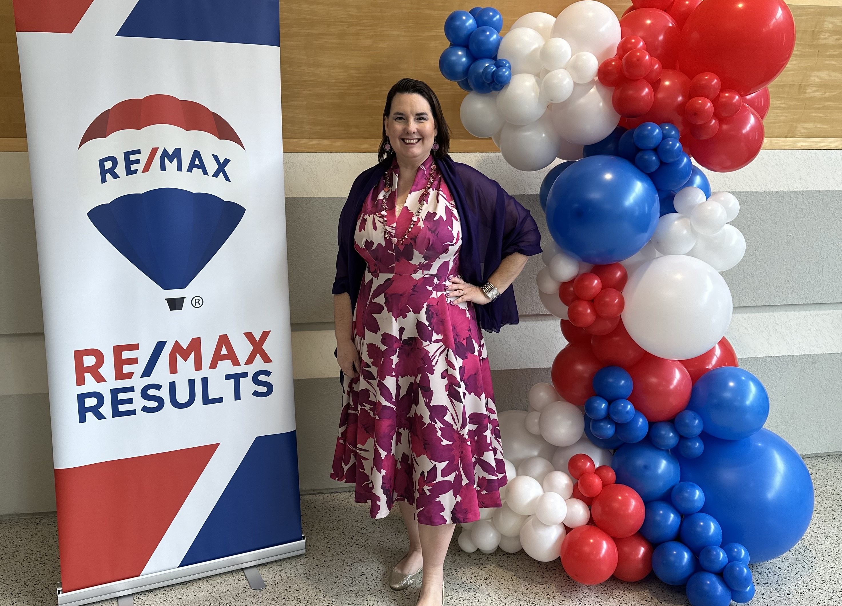 Lisa Collio smiling beside a RE/MAX Results banner at a company event, with a red, white, and blue balloon display.