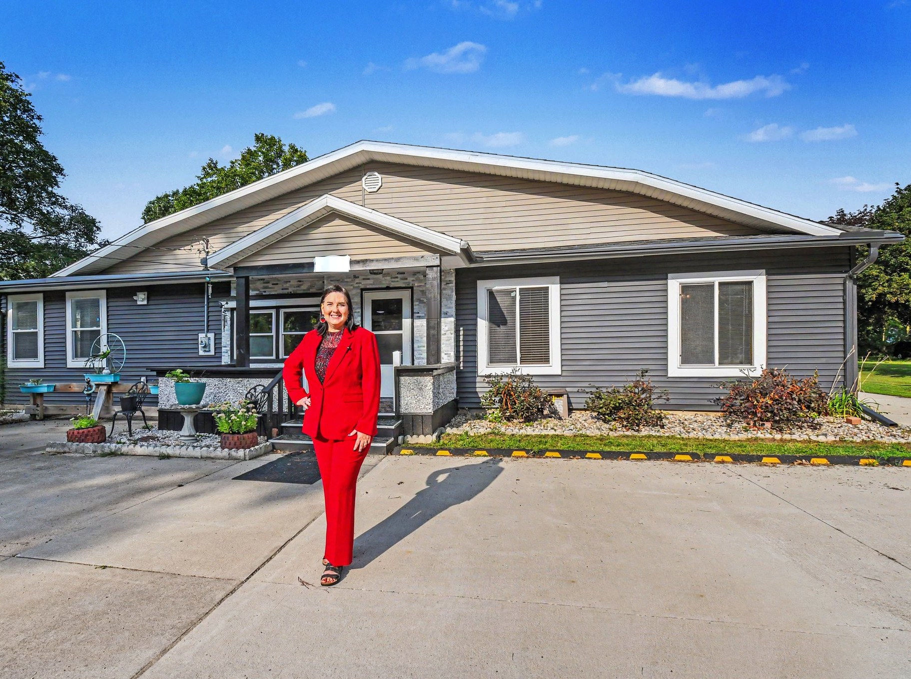 Lisa Collio, in a red suit, smiling in front of a gray single-story house with a stone-trimmed front porch on a sunny day.