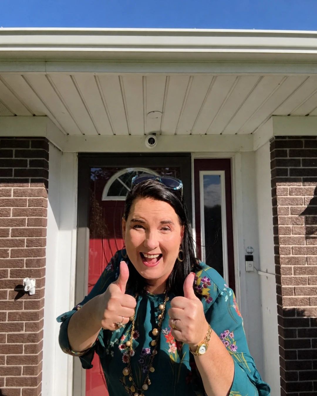 Lisa Collio smiling and giving two thumbs up in front of a red front door framed by brick columns.