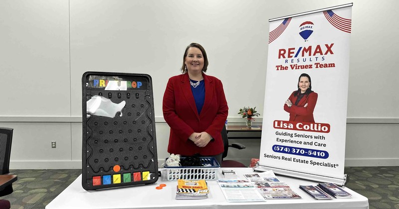 Lisa Collio at her RE/MAX Results, The Viruez Team information table at a seniors event, with a banner reading "Guiding Seniors with Experience and Care."