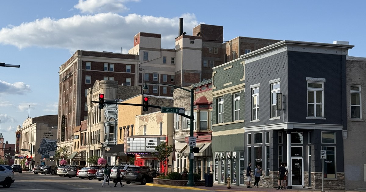 A downtown Elkhart block of two- and three-storey commercial buildings under a clear sky, with traffic signals over the street and people on the sidewalk.
