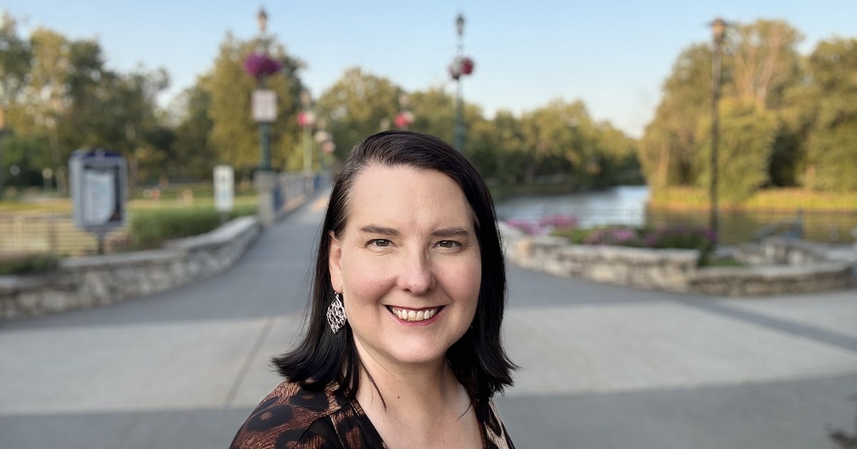 Lisa Collio smiling on a park path at golden hour, with a lamp-lined bridge and the river softly out of focus behind her.