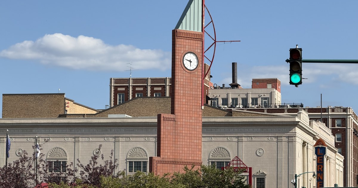 A brick clock tower rising above a pale stone civic building in downtown Elkhart, with a theatre's vertical blade sign on the building to its right.