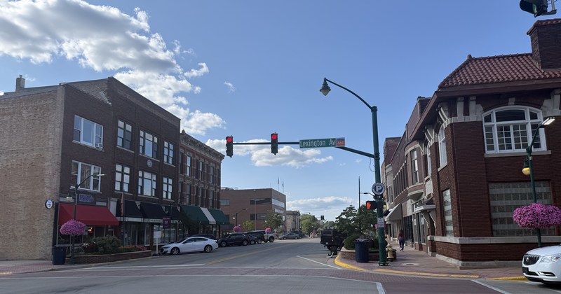 A downtown Elkhart intersection on a clear day, with brick commercial buildings on both sides of the street, awnings over the storefronts and planters of pink flowers at the corners.