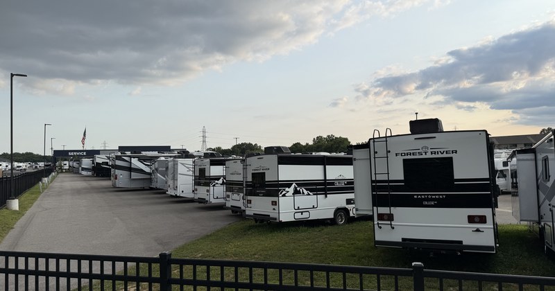 A row of new recreational vehicles lined up on a lot at dusk behind a metal fence, under a heavy sky.