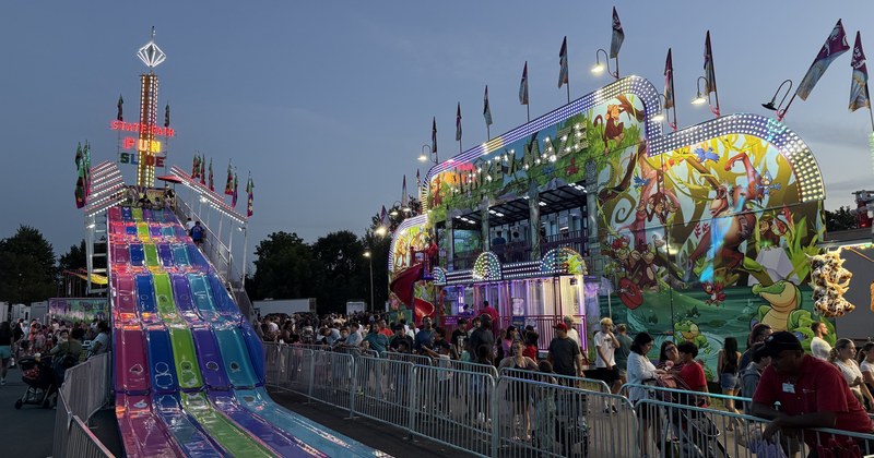 A fairground midway at dusk, with a brightly lit multi-lane slide on the left and a painted funhouse ride under strings of lights on the right.