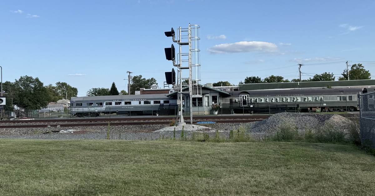 A silver passenger railcar lettered NEW YORK CENTRAL and CITY OF ELKHART standing on a siding, with a signal gantry and tracks in front of it.