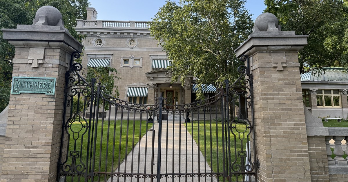 The stone gateposts and wrought-iron gate at the entrance to Ruthmere, with the mansion's front facade and striped awnings visible beyond the lawn. A bronze plaque reading RUTHMERE is mounted on the left post.