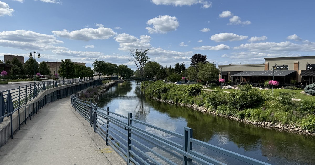 A riverwalk with a blue metal railing running beside the water, with a landscaped bank and low buildings along the far side.