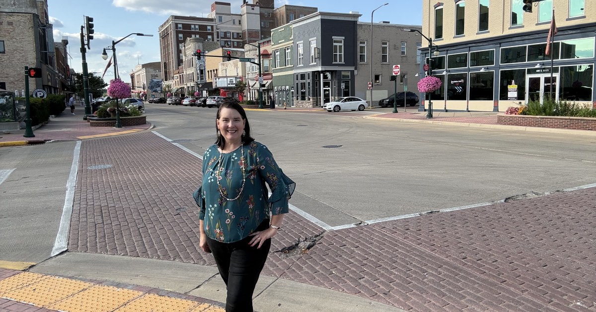 Lisa Collio standing at a brick crosswalk in downtown Elkhart, with storefronts and traffic signals along the street behind her.
