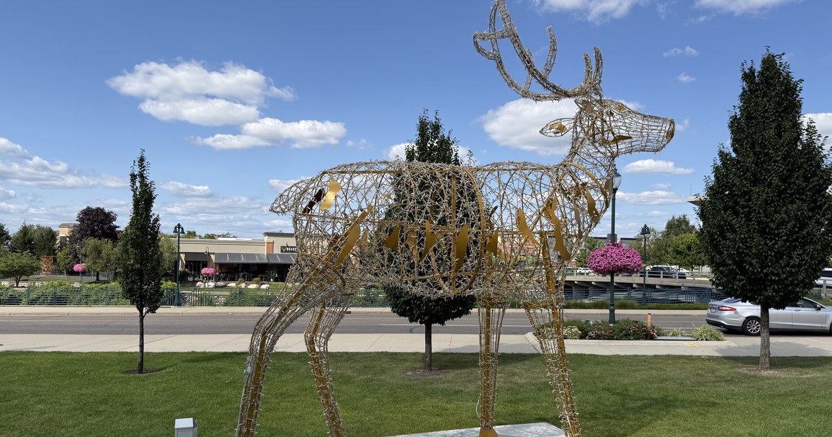 A large open-frame deer sculpture standing on a lawn beside a road, with trees and low buildings behind.