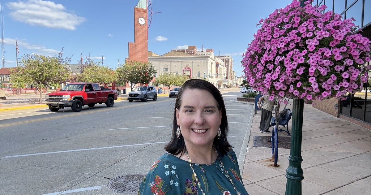 Lisa Collio smiling on a downtown Elkhart sidewalk, with a brick clock tower and a hanging basket of pink flowers behind her.