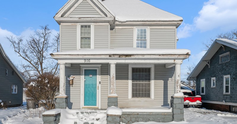 An older two-storey home with a covered front porch and a turquoise front door, with snow on the ground.