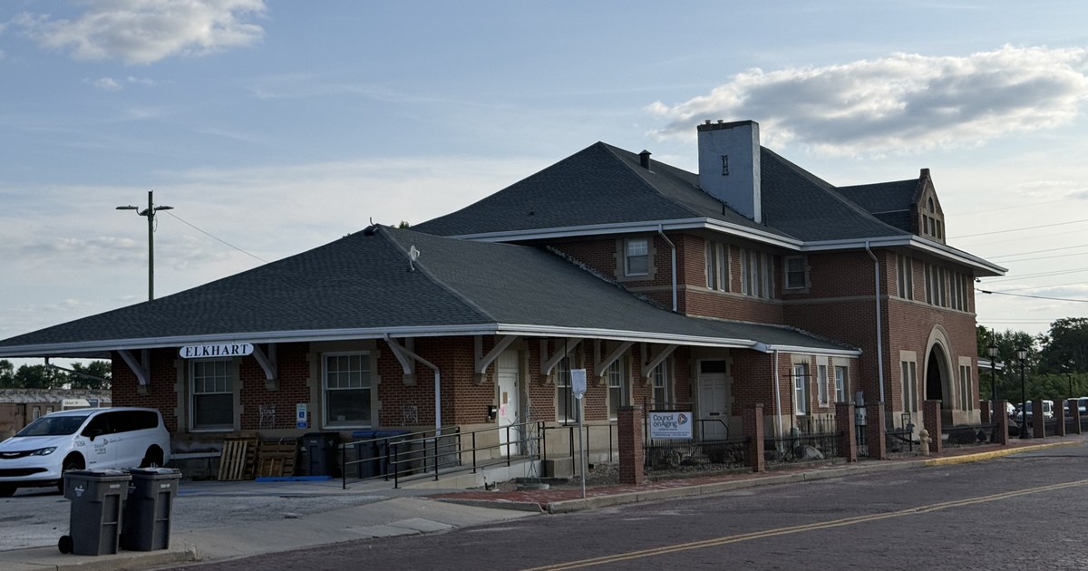 A single-storey brick railway station with a wide overhanging roof, the word ELKHART on the canopy above the platform, beside an empty street.