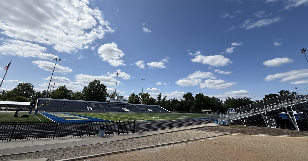 A school athletic field with tiered bleachers, floodlight towers and a running track under a partly clouded sky.