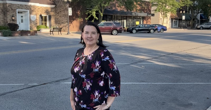 Lisa Collio standing in a downtown Goshen street, with brick storefronts, parked cars and street trees behind her.