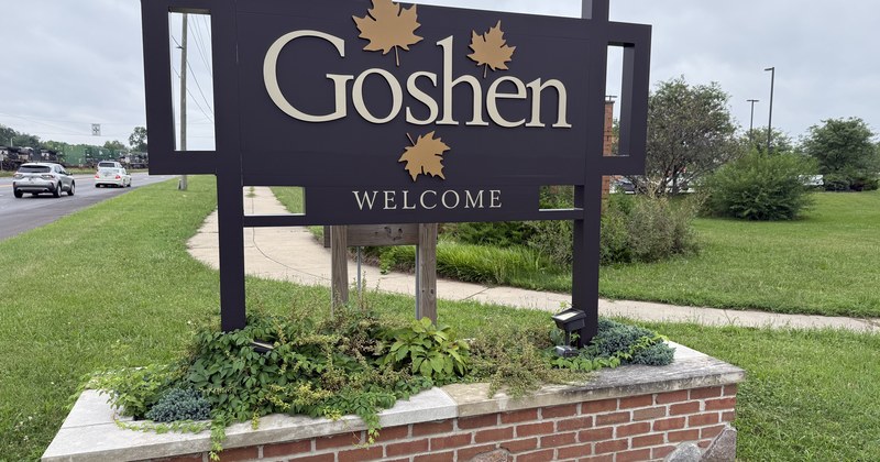 A wooden Goshen welcome sign with gold lettering and maple leaf accents, set in a brick planter.