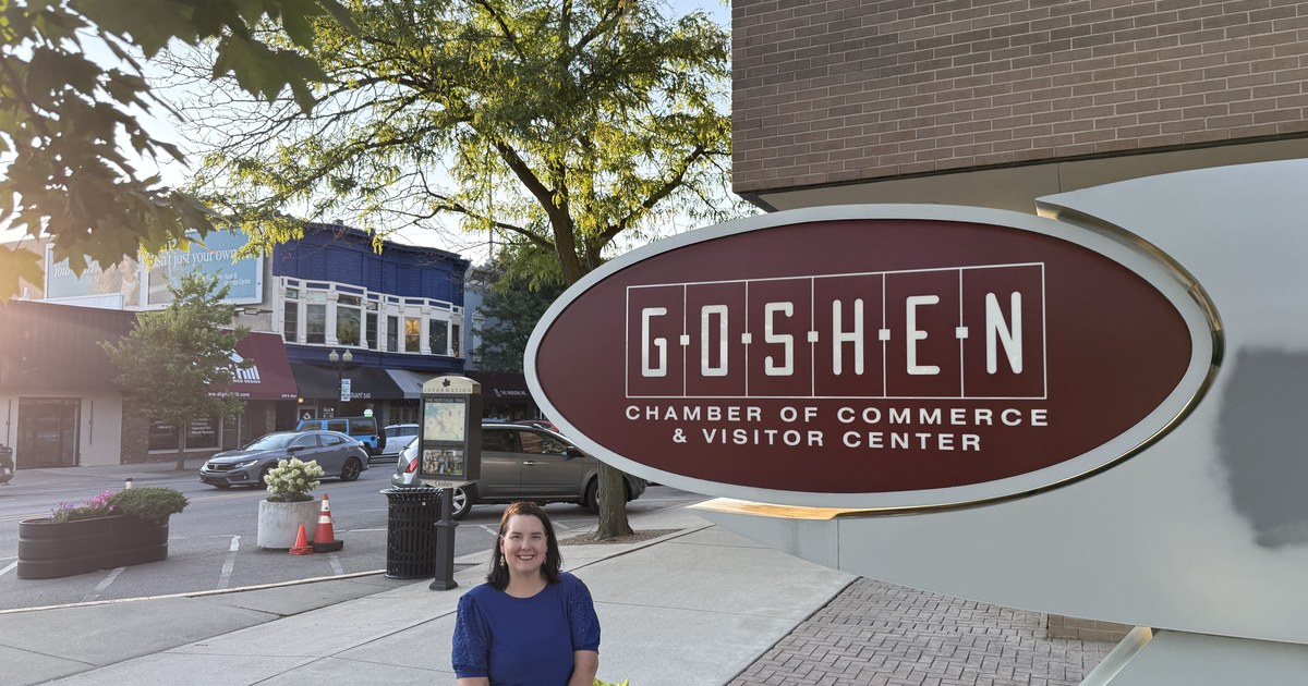 Lisa Collio standing beside a large oval sign mounted on a brick building in downtown Goshen.
