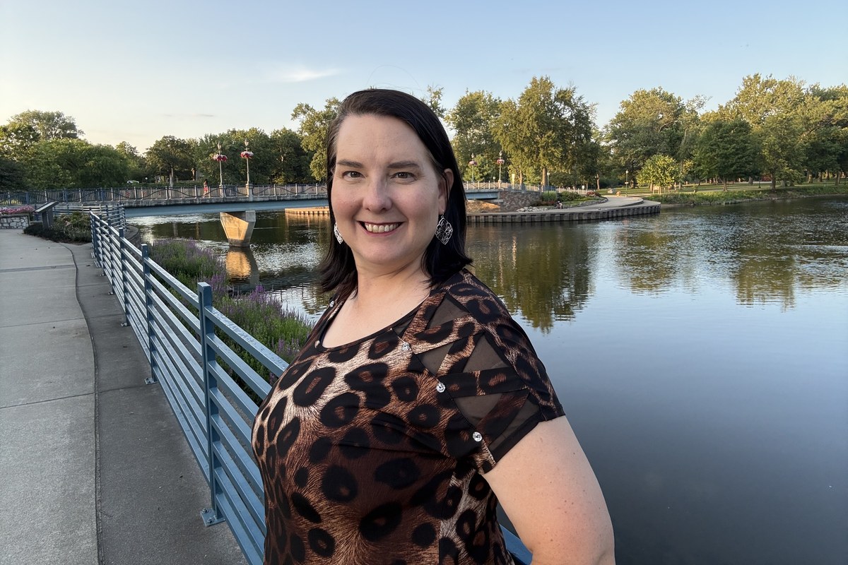 Lisa Collio standing beside the river in Elkhart, Indiana, next to a blue-railed bridge lined with lampposts and hanging flower baskets.