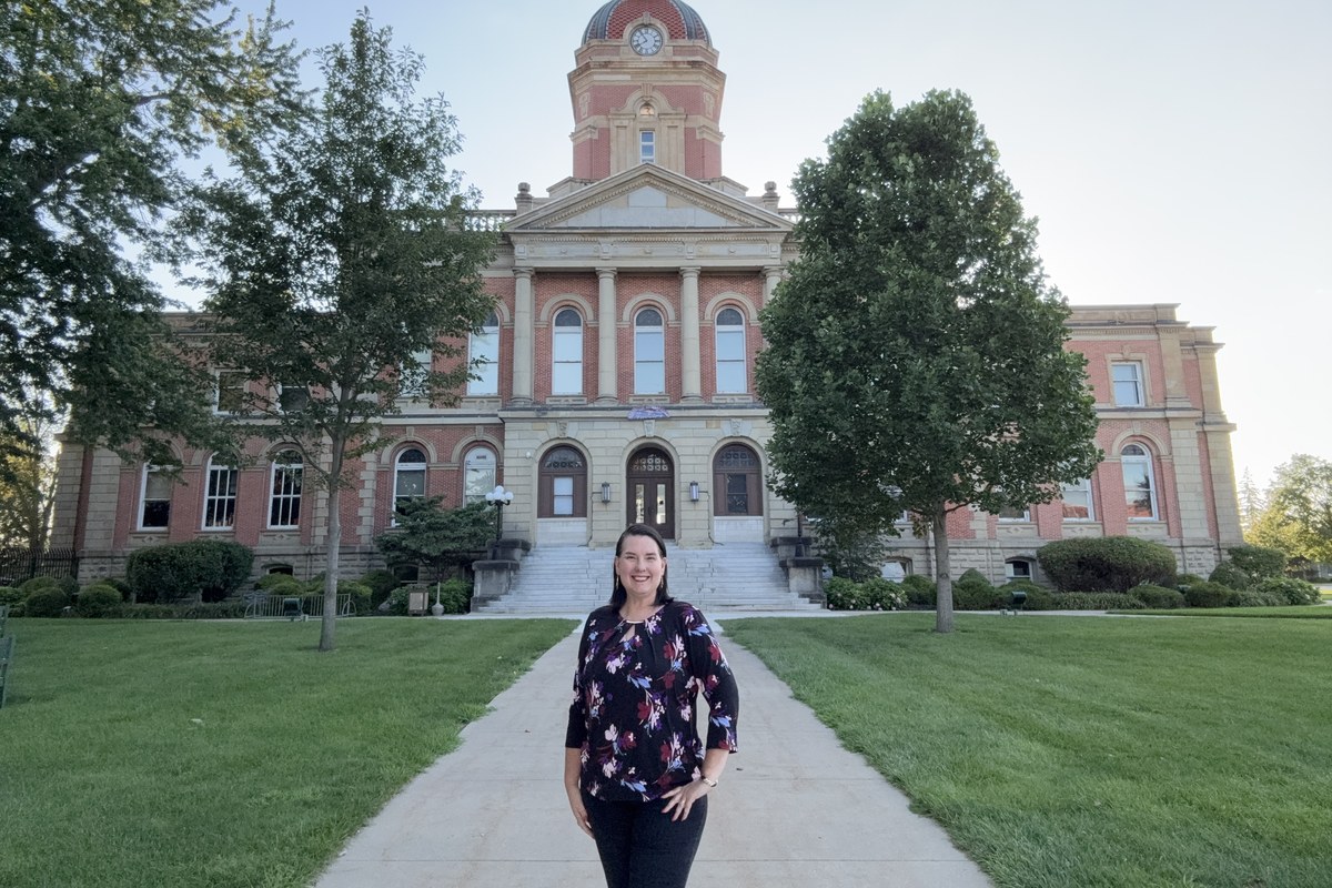 Lisa Collio standing on the lawn in front of the Elkhart County Courthouse in Goshen, Indiana, with its brick clock tower behind her.