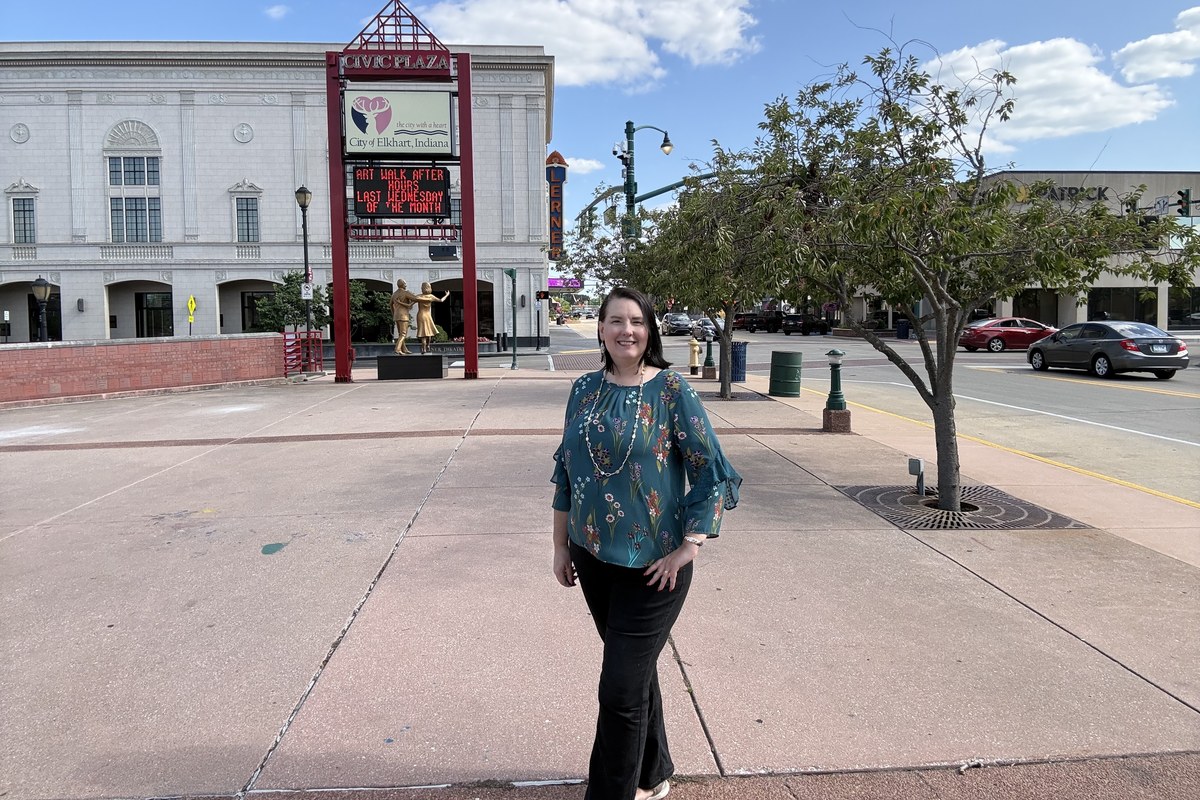 Lisa Collio standing in the civic plaza in downtown Elkhart, Indiana, with a limestone municipal building and a bronze statue of a dancing couple behind her.