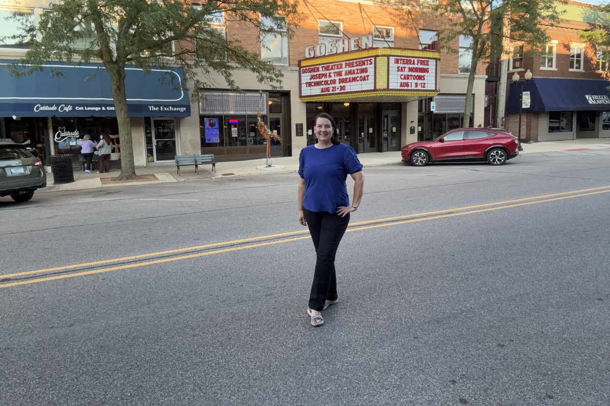 Lisa Collio standing on a downtown Goshen street in front of a historic theater marquee, with brick storefronts along the block behind her.